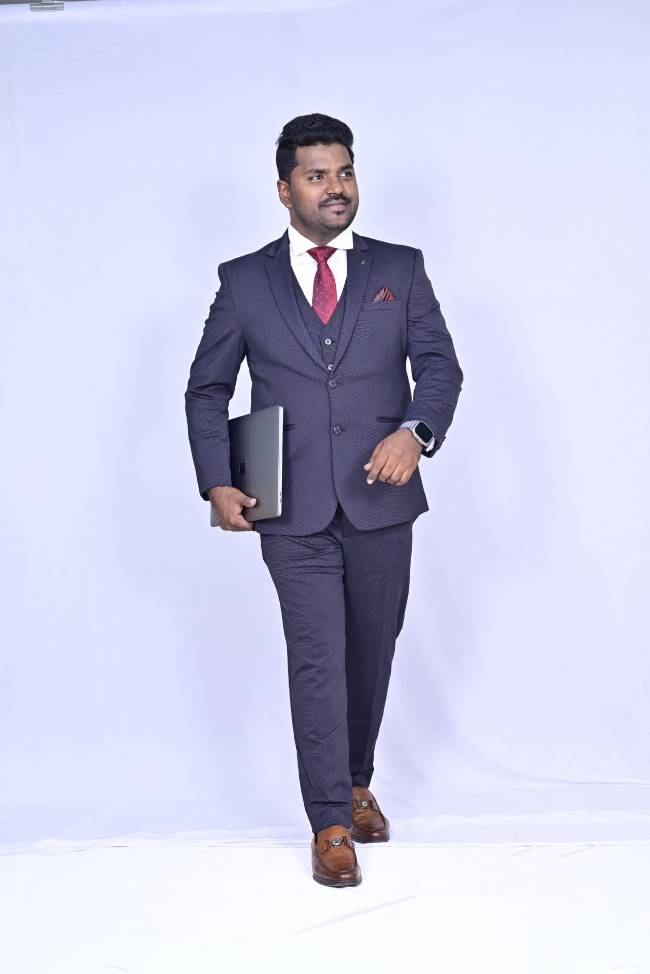 Dr. Ramesha A. walking with a laptop in a navy suit